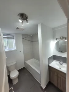 Salle de bain rénovée avec carrelage gris, luminaire moderne, et miroir élégant.