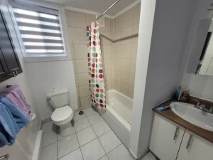 Salle de bain avant rénovation avec rideau de douche coloré, carrelage beige, et lumière naturelle.