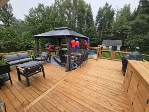 Grande terrasse en bois traité avec un gazebo décoré de ballons rouges et bleus, mobilier extérieur et vue sur la piscine